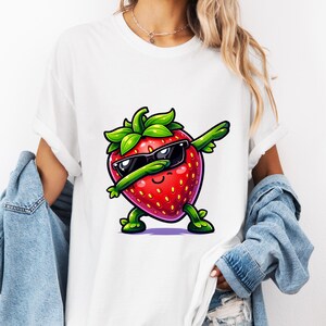 May include: White t-shirt featuring a cartoon strawberry wearing sunglasses and dabbing. The strawberry is red with green leaves and stems. The shirt is a casual style.