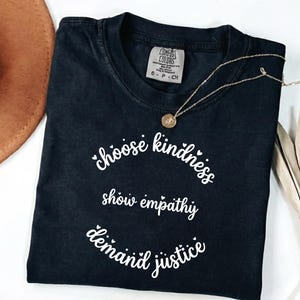Puede incluir: Camiseta negra con las palabras "choose kindness, show empathy, demand justice" en escritura blanca. Un collar dorado con un colgante descansa sobre la camiseta. Un sombrero marrón y una planta seca están en el fondo.