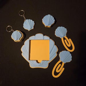 May include: A collection of seashell-themed desk accessories in blue and yellow. Includes a memo holder with a yellow sticky note, keychains, paper clips, and decorative shells. The items are arranged on a black surface.