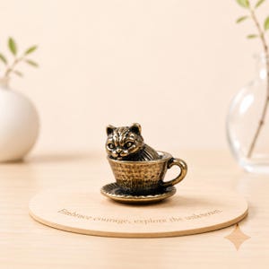 May include: A bronze-colored cat figurine sits inside a teacup on a saucer. The teacup and saucer are also bronze-colored. The cat is detailed with visible fur and facial features. The teacup is on a wooden coaster with the text "Embrace courage, explore the unknown."