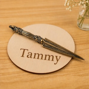 May include: A circular light-colored wooden coaster featuring the name Tammy engraved in a dark serif font. Resting diagonally across the coaster is an antique brass-toned metal letter opener with an ornate, textured handle and a pointed blade.