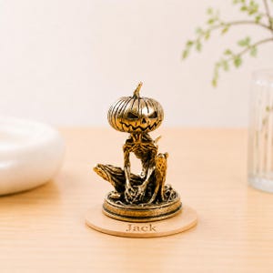 Könnte beinhalten: Goldfarbene Halloween-Figur mit einem Kürbiskopf mit geschnitzten Gesichtszügen auf einem skelettartigen Körper. Die Figur steht auf einem Holzsockel mit dem eingravierten Namen "Jack". Das Design hat ein gruseliges, festliches Thema.