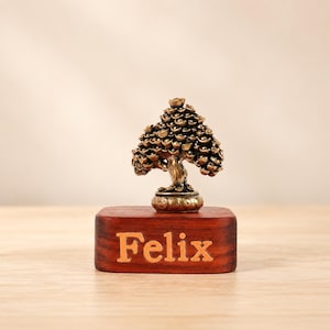 Könnte beinhalten: Eine bronzefarbene Metallskulptur eines Baumes mit runden, münzähnlichen Blättern auf einem rechteckigen Holzsockel. Auf der Vorderseite des dunklen Holzsockels ist der Name Felix in einer hellen, serifenlosen Schrift eingraviert.