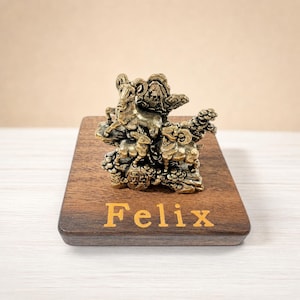 Könnte beinhalten: Eine messingfarbene Metallskulptur mit drei Widdern auf einem felsigen Sockel, montiert auf einem rechteckigen dunklen Holzblock mit dem Namen Felix, der in goldfarbenen Buchstaben auf der Vorderseite des Holzsockels eingraviert ist.