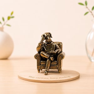 May include: A bronze-effect dog figurine seated in an armchair, reading a book. The figurine is on a wooden coaster with the inscription "Stay curious, keep reading."