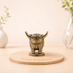 Könnte beinhalten: Eine bronzefarbene Highland-Kuh-Figur mit großen Hörnern, die auf einem runden Holzsockel steht. Auf dem Sockel steht der Text "Fortune & Resilience". Es ist ein dekorativer Artikel.