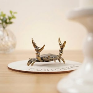 Op de afbeelding: Een bronzen krabfiguur met omhoog geheven scharen, rustend op een ronde houten onderzetter. De onderzetter heeft het woord "CURIOSITY" erop gedrukt. De krab is gedetailleerd en lijkt van metaal te zijn.