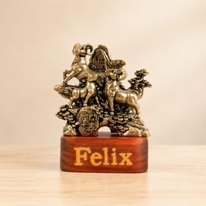 Könnte beinhalten: Eine messingfarbene Metallskulptur mit drei Widdern auf einer felsigen Landschaft mit eingravierten chinesischen Schriftzeichen, montiert auf einem rechteckigen dunklen Holzsockel mit dem Namen Felix in goldener Schrift.