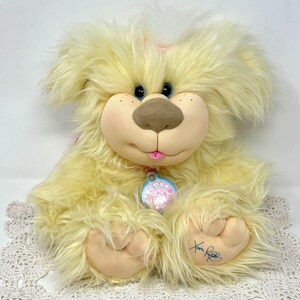 May include: A plush, yellow dog with a soft, fluffy texture. The dog has a light brown nose, blue eyes, and a pink tongue. It wears a pink collar with a tag. The dog is sitting on a white lace surface.
