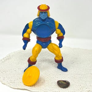 Puede incluir: Figura de acción vintage de un personaje musculoso en azul, amarillo y rojo. La figura tiene una cara y un casco azules, brazos y piernas amarillos y detalles en rojo. Un disco amarillo y un pequeño objeto de color oscuro están cerca de la figura.