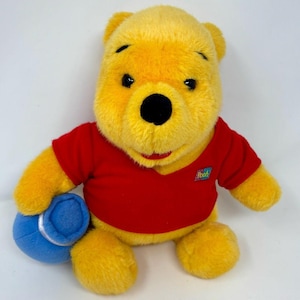 Könnte beinhalten: Ein Winnie Puuh Plüschtier mit gelbem Fell, einer schwarzen Nase und einem roten T-Shirt. Das T-Shirt hat einen kleinen rechteckigen Aufnäher mit dem Text Pooh. Der Bär hält in seiner rechten Hand einen blauen, zylindrischen Topf mit silbernem Rand.