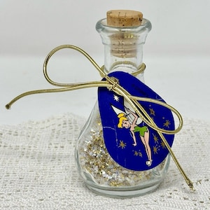 Puede incluir: Una botella de vidrio transparente con tapón de corcho, llena de purpurina dorada. Una etiqueta azul con una ilustración de Campanilla está sujeta con hilo dorado. La botella está atada con un cordón dorado.