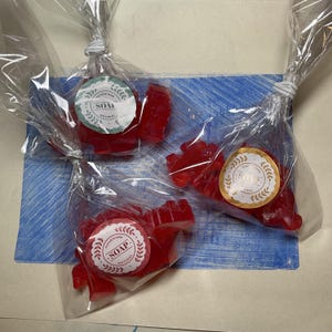 May include: Three handmade red soaps, shaped like dinosaurs, are individually wrapped in clear cellophane. Each soap has a round sticker that reads "handmade SOAP natural". The soaps are tied with a white twist tie.
