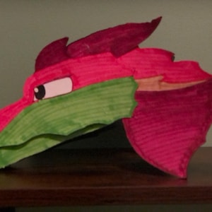 Può includere: Una maschera a forma di testa di drago fatta a mano, principalmente rosa e verde, con un design a cartone animato. La testa del drago presenta una cresta rosa, una mascella verde e un occhio bianco e nero. La maschera è probabilmente fatta di carta o cartone.