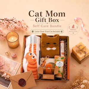 K&ouml;nnte beinhalten: Eine Geschenkbox mit einem Paar wei&szlig;er Socken mit orangefarbenem Katzenmotiv, braunen Katzensocken, einer Melonen-Hand- und K&ouml;rpercreme, einem Lippenbalsam, einer kleinen Katzenfigur, einem Schl&uuml;sselanh&auml;nger und einem braunen Umschlag mit Wachssiegel.
