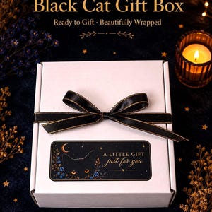 K&ouml;nnte beinhalten: Eine wei&szlig;e, rechteckige Geschenkbox, die mit einem schwarzen Band mit goldenem Rand verschlossen ist. Ein rechteckiges Etikett auf der Vorderseite zeigt die Silhouette einer schwarzen Katze mit leuchtend orangen Augen, blauen Blumen, einem Halbmond und dem Text A LITTLE GIFT just for you.