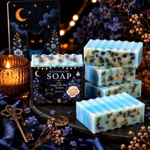 K&ouml;nnte beinhalten: Vier rechteckige St&uuml;cke hellblauer handgemachter Seife mit eingearbeiteten getrockneten Kr&auml;utern und Goldflocken, gestapelt neben einer schwarzen Schachtel mit der Aufschrift HANDMADE SOAP with Forget-Me-Not & Rose, verziert mit einer schwarzen Katze.