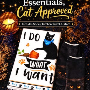 K&ouml;nnte beinhalten: Ein wei&szlig;es K&uuml;chentuch mit einem schwarzen Katzenmotiv, blauem Halstuch, einem umgekippten orangefarbenen Topf und dem Text I DO WHAT I WANT, dazu ein Paar schwarze Socken mit einem realistischen Katzenkopf-Druck und leuchtend orangen Augen.