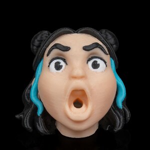 May include: A 3D-printed figurine of a surprised face with wide eyes and an open mouth. The figure has black hair styled in two buns, with blue accents. The skin tone is a light peach color, and the background is black.
