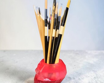 May include: A collection of paint brushes in a red skull-shaped holder. The brushes have black and tan handles with clear plastic tips. The holder is made of a translucent red material.