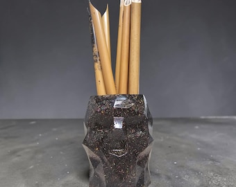 May include: A dark, faceted, geometric pen holder with multicolored glitter accents containing several light brown bamboo calligraphy pens with pointed nibs. The container has a textured, multi-angled surface and a deep, dark finish.