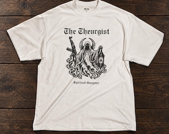 May include: An off-white short-sleeve t-shirt featuring a black graphic print of a hooded figure holding a rifle and a lantern. The text "The Theurgist" appears above the figure, and the words "Spiritual Gangster" are printed below the graphic.