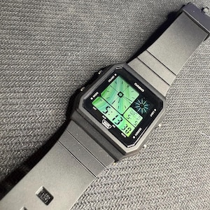 May include: A gray digital wristwatch with a square face and a matching gray band. The watch face displays the time, day, and date in a green hue. The brand name "CASIO" is visible on the watch face.