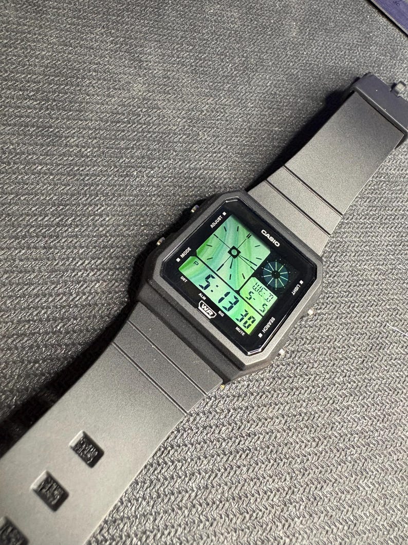 May include: A gray digital wristwatch with a square face and a matching gray band. The watch face displays the time, day, and date in a green hue. The brand name "CASIO" is visible on the watch face.