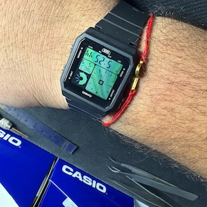 May include: Black digital Casio watch displaying the time 5:25:44. The watch has a rectangular face with a black strap. The watch is on a wrist with a red string bracelet. The word "CASIO" is printed on the watch face.