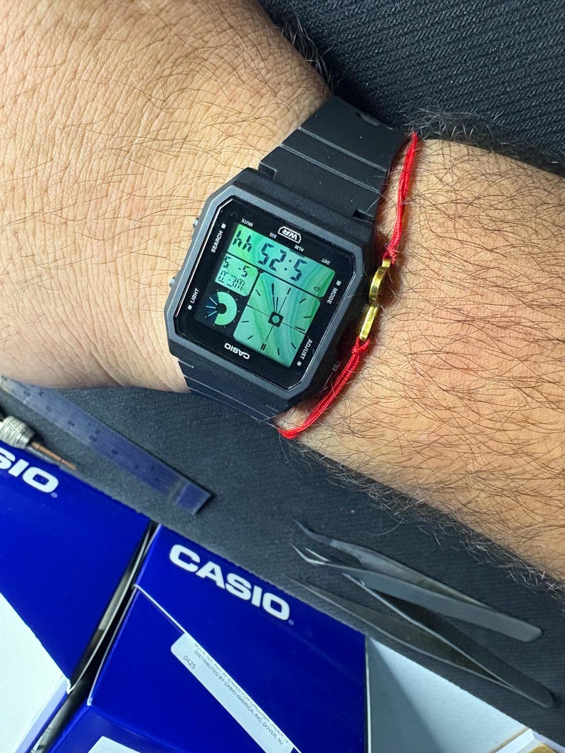 May include: Black digital Casio watch displaying the time 5:25:44. The watch has a rectangular face with a black strap. The watch is on a wrist with a red string bracelet. The word "CASIO" is printed on the watch face.