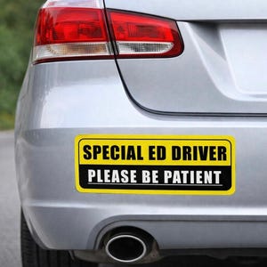 May include: A yellow and black bumper sticker on the back of a silver car. The sticker reads "SPECIAL ED DRIVER PLEASE BE PATIENT" in bold white text.