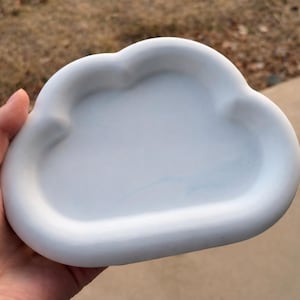 May include: A white, cloud-shaped ceramic tray. The tray has a smooth, rounded design and is held in a hand. The background is blurred, with a mix of brown and tan colors.