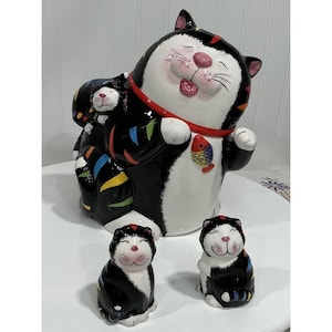 Può includere: Un set di statuine di gatti in ceramica con un grande gatto bianco e nero con un collare rosso e un gatto piccolo sulla schiena. Sono presenti anche due statuine di gatti più piccole. I gatti hanno accenti colorati e un dettaglio di pesce.