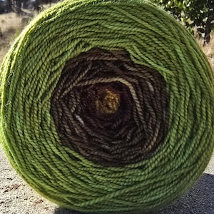 May include: A large ball of yarn with a gradient of colors, transitioning from a bright green exterior to a dark brown center. The yarn is tightly wound, creating a circular shape. The ball sits on a concrete surface, with a natural background.