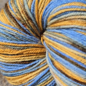 May include: Close-up of a ball of yarn with a mix of colors. The yarn features a blend of blue, gold, brown, and green hues. The yarn is tightly wound, showcasing the texture and color variations. Ideal for knitting and crochet projects.