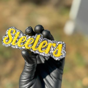Puede incluir: Logotipo rectangular y brillante de los Steelers en escritura amarilla sobre un fondo plateado y negro. El logotipo est&aacute; sostenido por una mano enguantada contra un fondo borroso al aire libre. La palabra "Steelers" se muestra de forma destacada.
