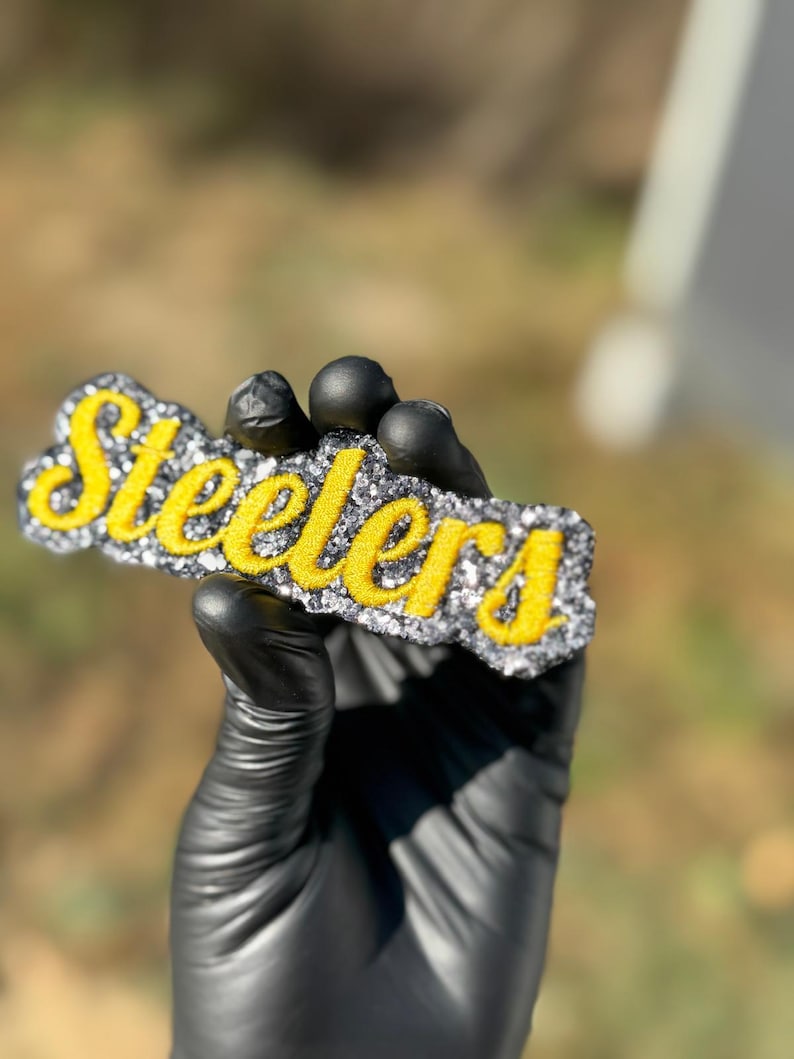 Puede incluir: Logotipo rectangular y brillante de los Steelers en escritura amarilla sobre un fondo plateado y negro. El logotipo est&aacute; sostenido por una mano enguantada contra un fondo borroso al aire libre. La palabra "Steelers" se muestra de forma destacada.