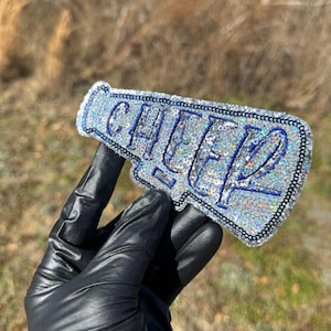 May include: A silver glitter megaphone-shaped keyring with the word "CHEER" in blue lettering. The keyring has a black border. The background is a blurred outdoor scene.
