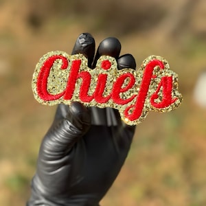 May include: A gold glitter patch with the word "Chiefs" in red, embroidered lettering. The patch is held up by a gloved hand against a blurred outdoor background.