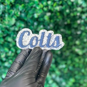 May include: A decorative patch featuring the word Colts in dark blue cursive embroidery. The lettering is outlined and set against a textured, shimmering silver glitter background, creating a distinct contrast between the blue text and silver base.