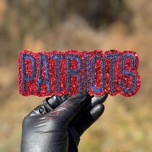 Könnte beinhalten: Ein rotes, glitzerndes "PATRIOTS"-Emblem mit blauen Buchstaben wird von einer behandschuhten Hand gehalten. Das Emblem hat ein strukturiertes, funkelndes Aussehen, und der Hintergrund ist unscharf.