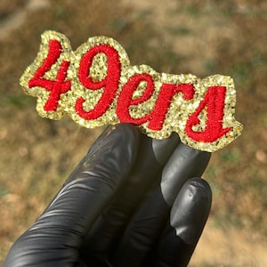 Peut inclure: Un écusson doré scintillant avec le mot "49ers" en rouge, tenu par une main gantée de noir. L'écusson est de forme rectangulaire aux bords arrondis. L'arrière-plan est flou, mettant en valeur les détails.