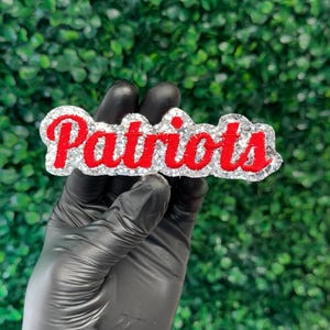 以下が含まれることがあります： キラキラ光る、シルバーと赤の「Patriots」エンブレム。「Patriots」の文字は赤色で、シルバーのグリッターで縁取られています。エンブレムは、緑色のテクスチャード加工された背景に対して、手袋をはめた手で持たれています。