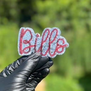 Puede incluir: Un parche decorativo con la palabra Bills escrita en lentejuelas rojas. Las letras están sobre un fondo de purpurina plateada con una forma troquelada que sigue el contorno del texto, creando una apariencia texturizada y reflectante.