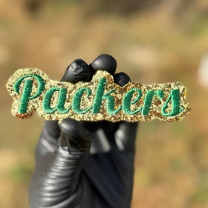 May include: A gold glitter "Packers" emblem with green lettering. The emblem is held by a person wearing a black glove. The background is blurred, with natural light.