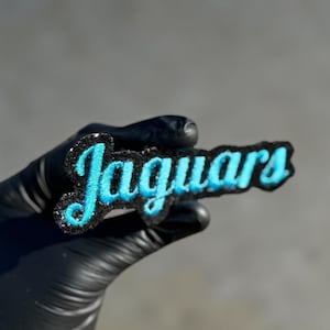 Może przedstawiać: Czarna i turkusowa naszywka z napisem "Jaguars" w ozdobnej czcionce. Naszywka ma czarną, błyszczącą obwódkę i jest trzymana przez dłoń w rękawicy.
