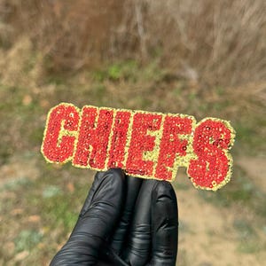 May include: A rectangular patch featuring the word CHIEFS in bold, block lettering. The letters are filled with vibrant red sequins, while the background of the patch is covered in shimmering gold sequins, creating a high-contrast, textured appearance.