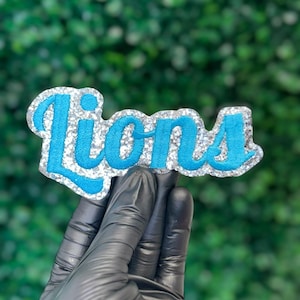 May include: A patch with the word "Lions" in turquoise script lettering, outlined in silver glitter. The patch is held by a gloved hand against a green, leafy background. The patch is likely for apparel.