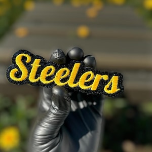 May include: An embroidered patch featuring the word "Steelers" in yellow script on a black background. The patch is held by a gloved hand. The background is blurred with green and yellow, suggesting an outdoor setting.