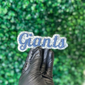 Op de afbeelding: Een glinsterend, marineblauw en zilver "Giants" embleem. Het woord "Giants" staat in een cursief lettertype, met een zilveren glitterrand. Het embleem wordt vastgehouden door een gehandschoende hand tegen een groene, bladerrijke achtergrond.